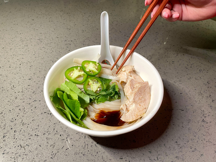 A white bowl sits center on a gray granite countertop with a white soup spoon resting on the far edge. Inside the bowl is Justin's homemade Pho. A hand holding wooden chopsticks hovers above the dish.