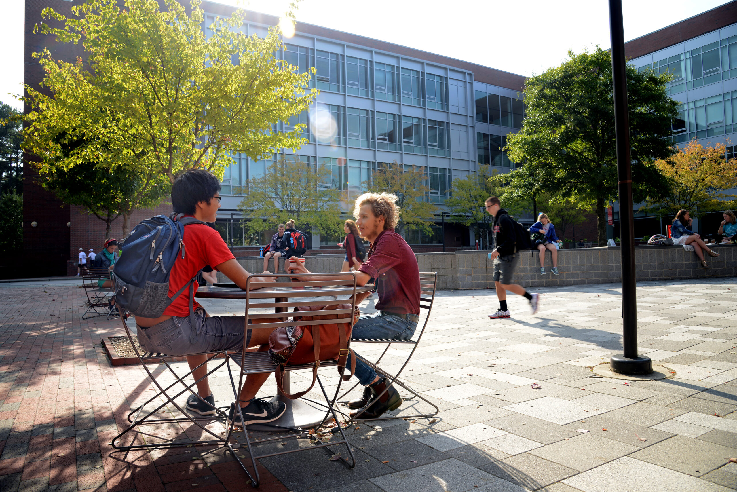 Two students sitting at a table outdoors in the foreground in front of SAS hall with other students doing other morning class activities.