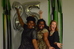 Three women pose in front of silver balloons and green streamers at photo booth