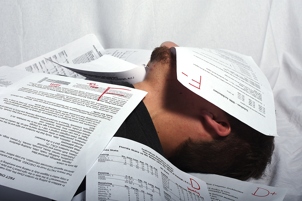 A student lays buried under a pile of white papers. The papers have big red letters on them ("F" and "D+") that show the student is failing their courses.
