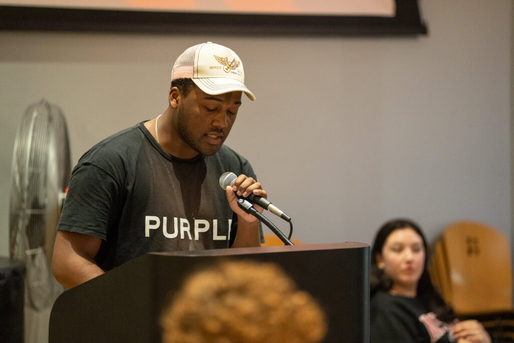 Image of a speaker in a baseball cap and t shirt reading his literary piece to a crowd.